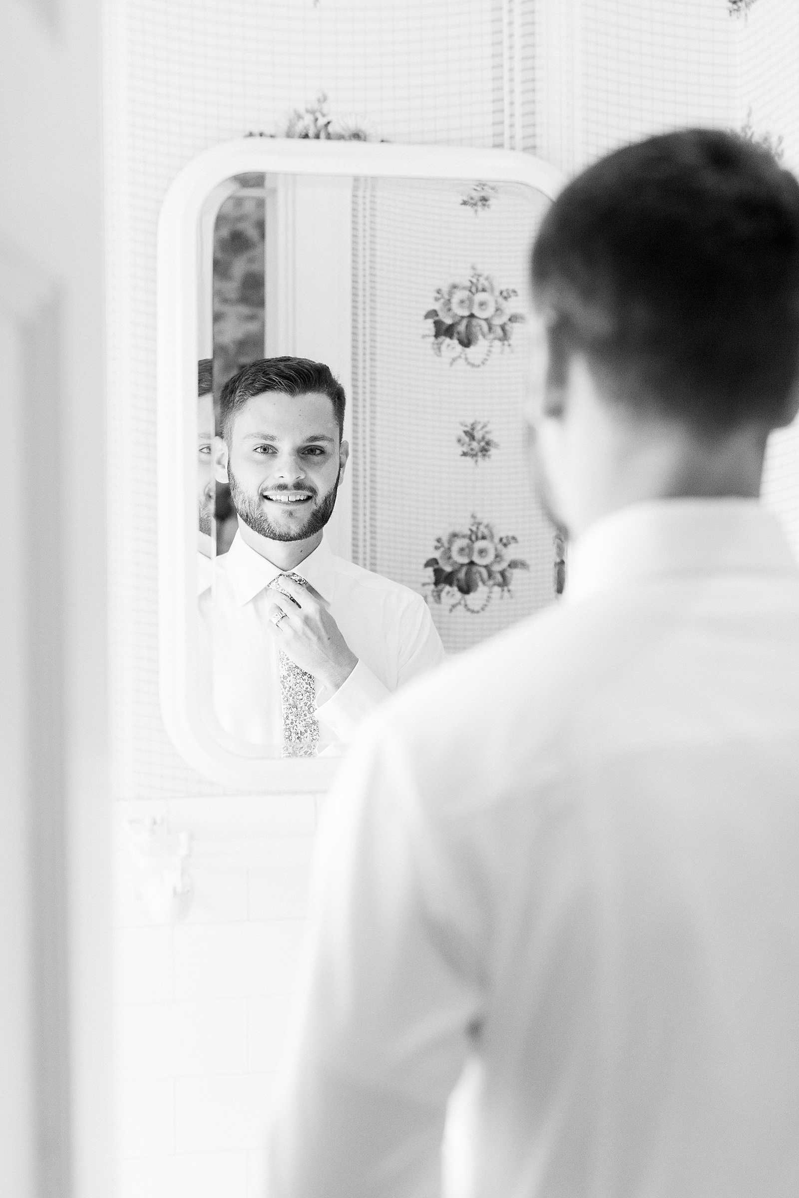 Black and white image of groom looking into a mirror and adjusting his tie.