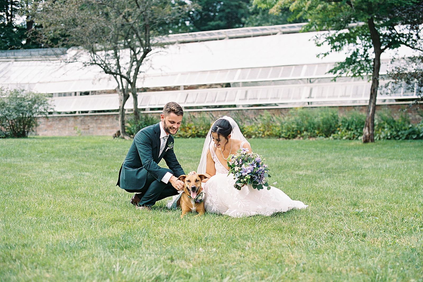 Bride and groom kneeling on a garden lawn to play with their dog. 