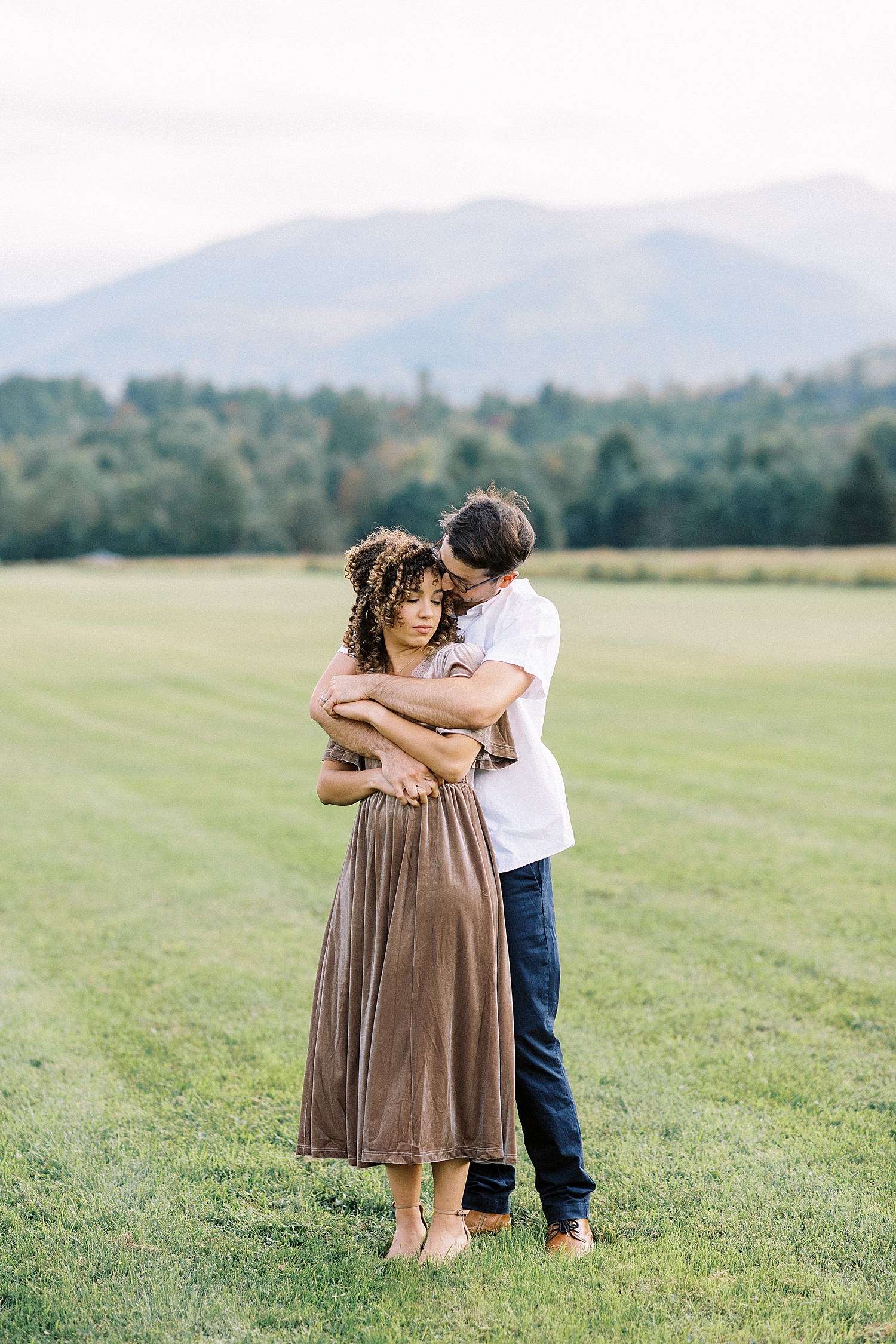 Man embracing his wife in a field with mountains behind them, shot by New York Photographer, Lynne Reznick.