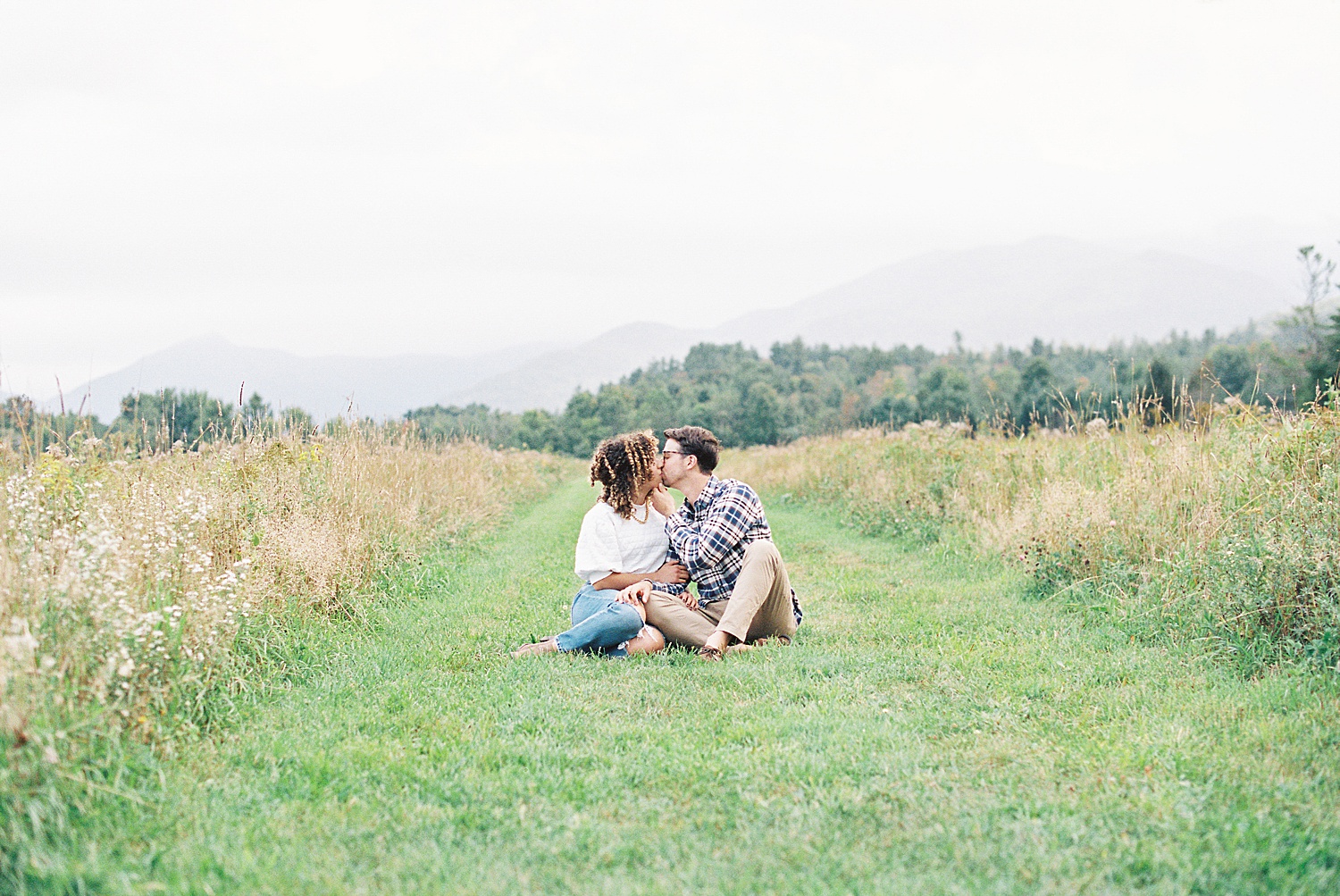 Married couple kissing in a field with the mountains in the foggy background.