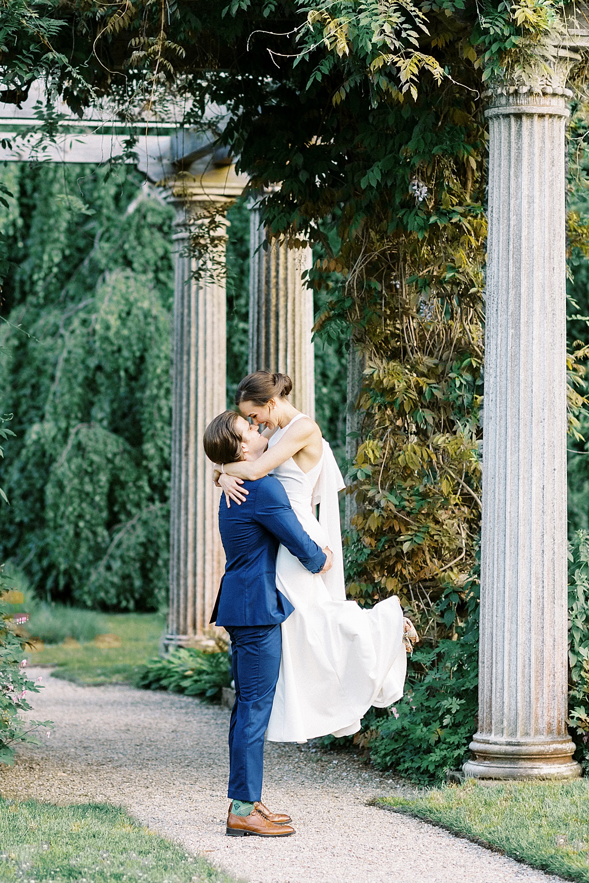 groom in blue suit sweeps bride off her feet beneath columns in garden venue shot by Massachusetts wedding photographer