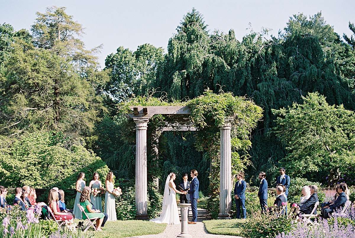 bride and groom stand beneath elegant columns in garden venue during ceremony shot by Massachusetts wedding photographer