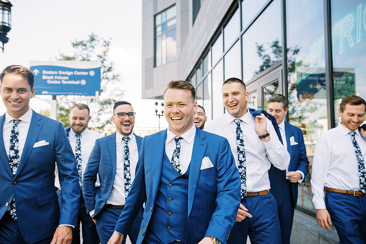 groom in three piece blue suit and groomsmen in floral ties walk pier before nautical oceanview wedding