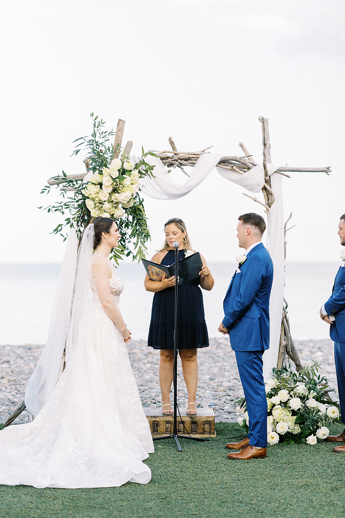 Bride in lace gown and groom in blue suit share vows during ocean side wedding shot by Lynne Reznick Photography