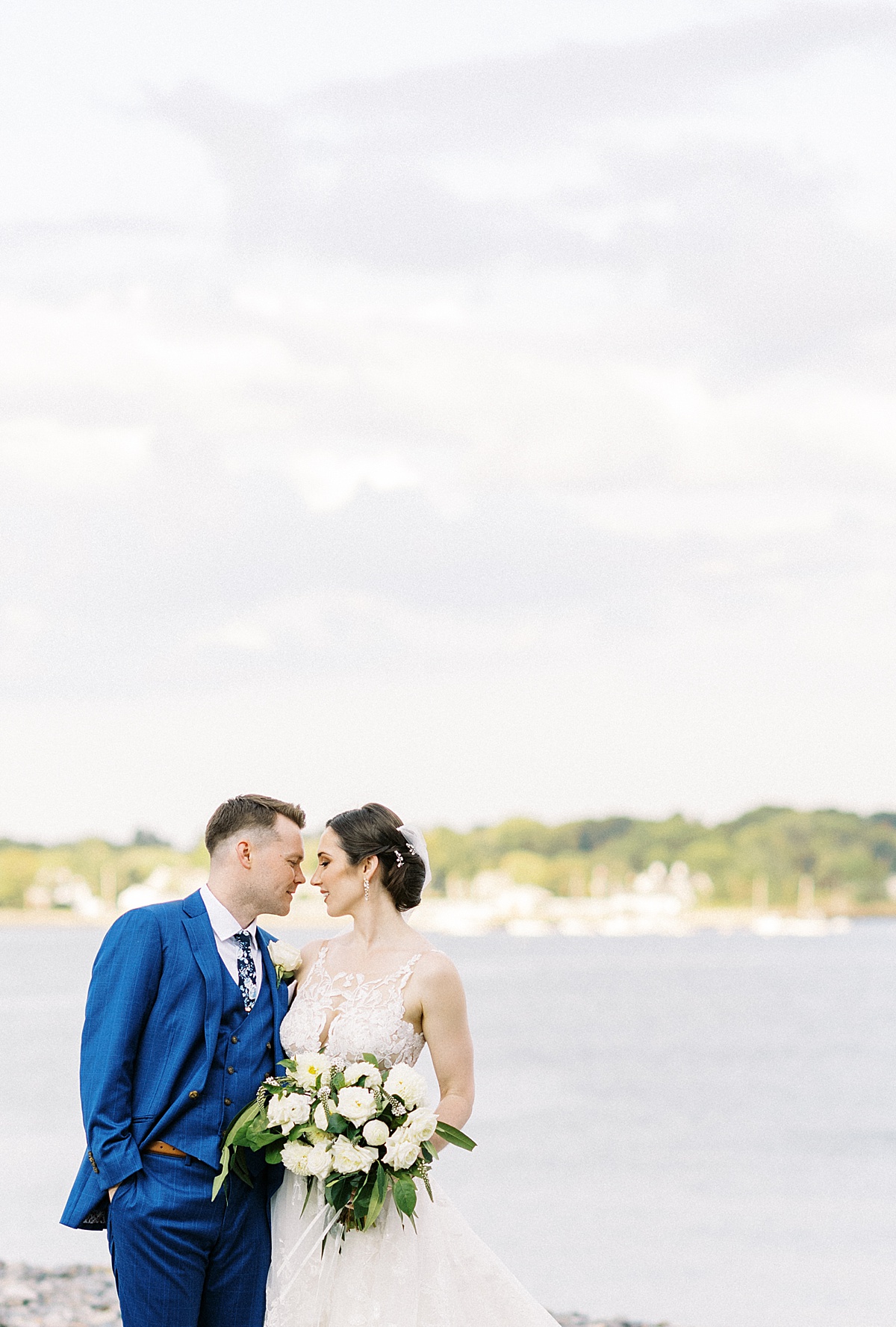 classy bride and groom in white lace and blue tux pose by water shot by Lynne Reznick Photography