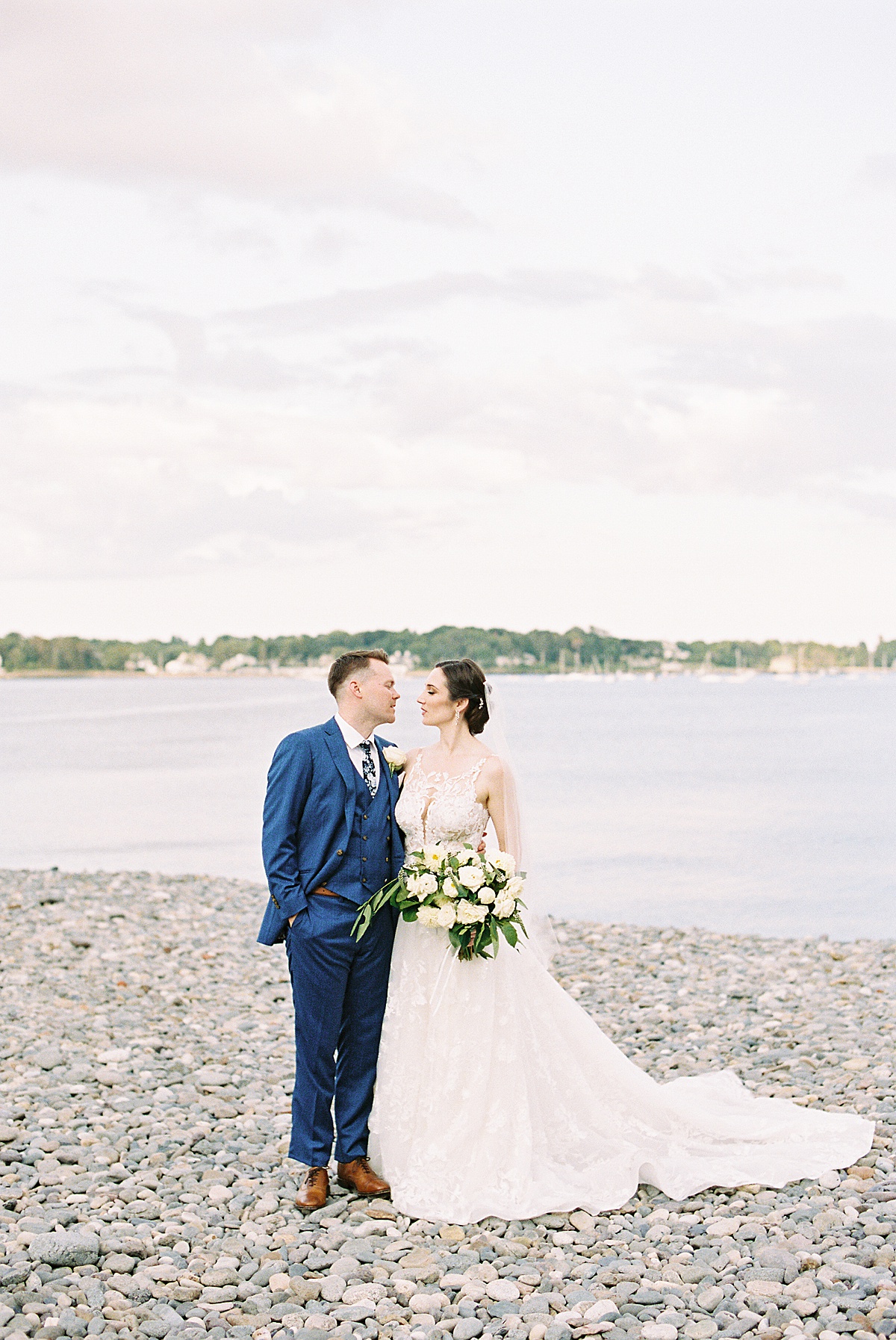 bride in romantic gown and groom in preppy blue suit pose on pebble beach for Massachusetts wedding photographer
