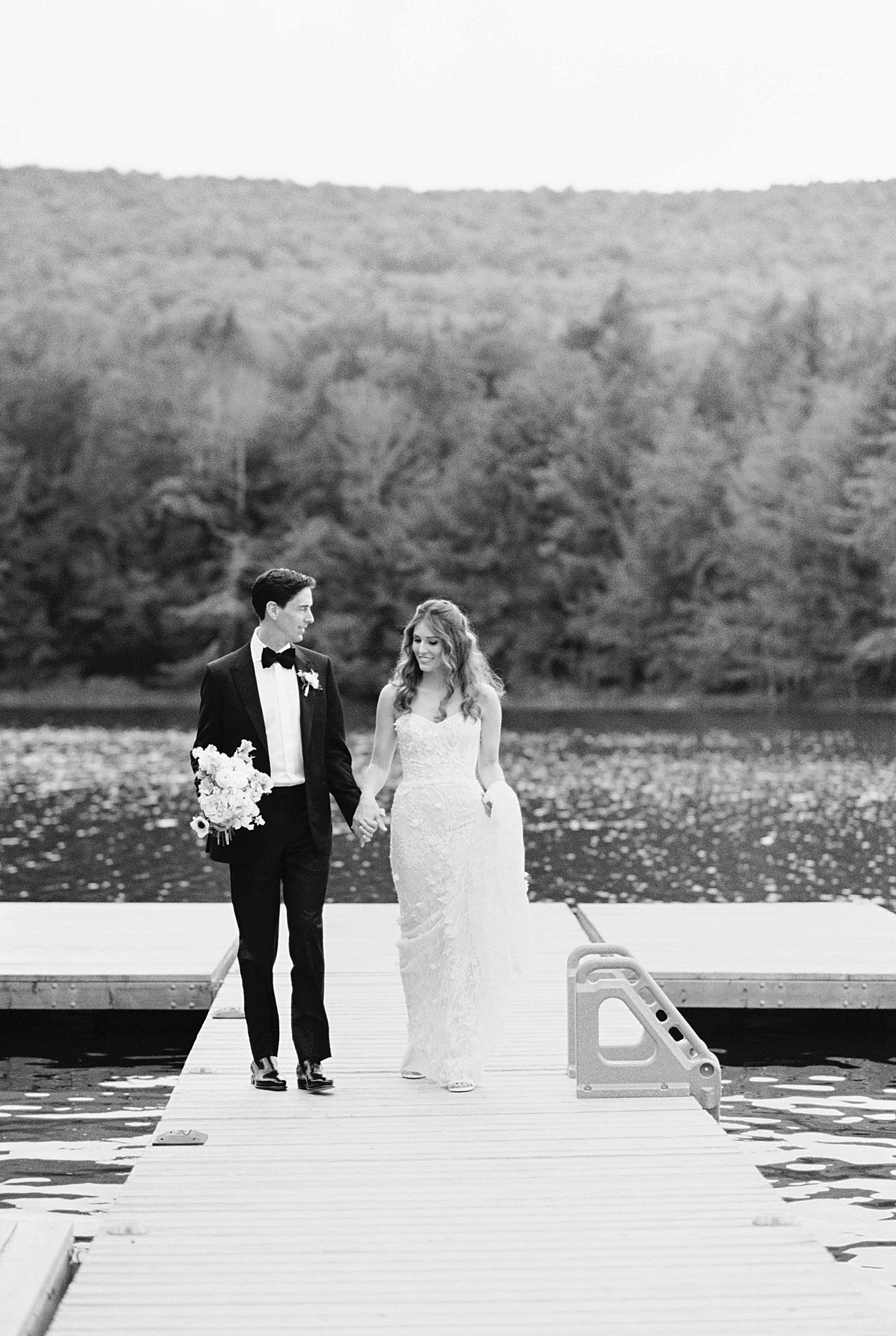 newlyweds hold hands as they walk on dock at lake in Catskills by Lynne Reznick Photography 