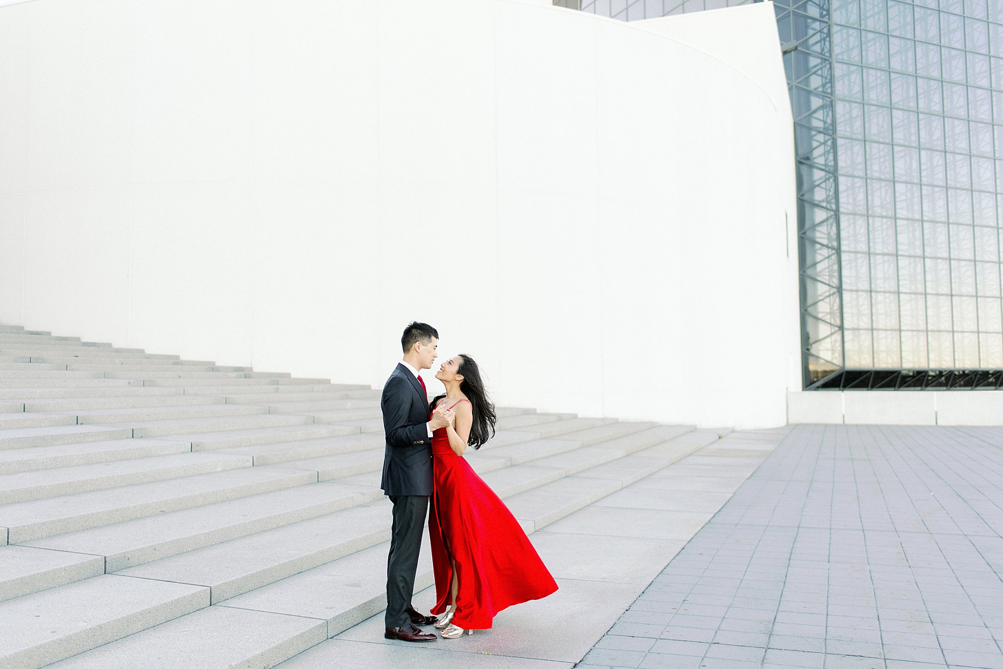 couple looks at each other as her red dress is blown by wind by Boston family portrait photographer