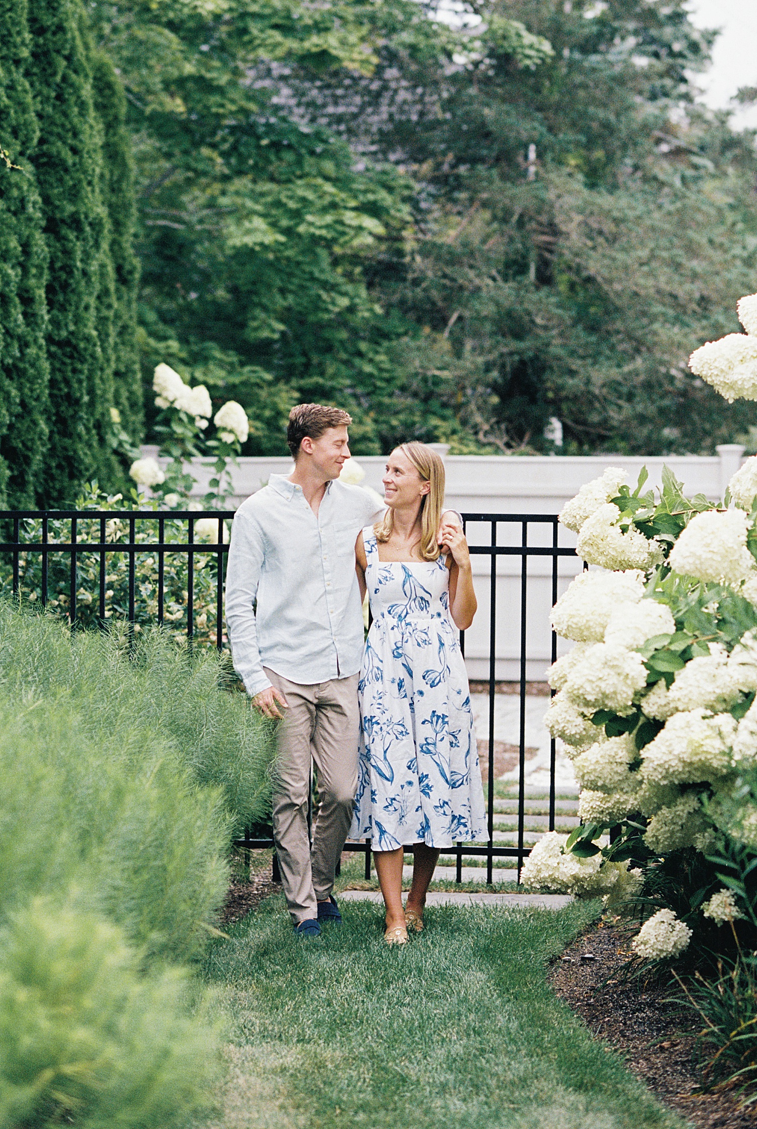 blonde woman in white & blue dress walks with fiancé in garden in Cotuit Bay