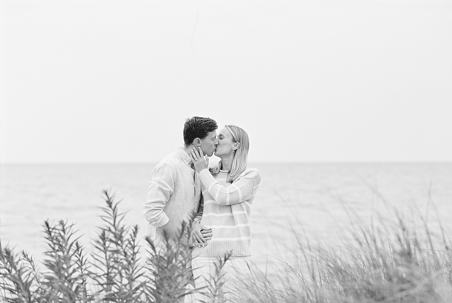 Woman leans over to kiss her fiancé by the ocean by Cape Cod Wedding Photographer