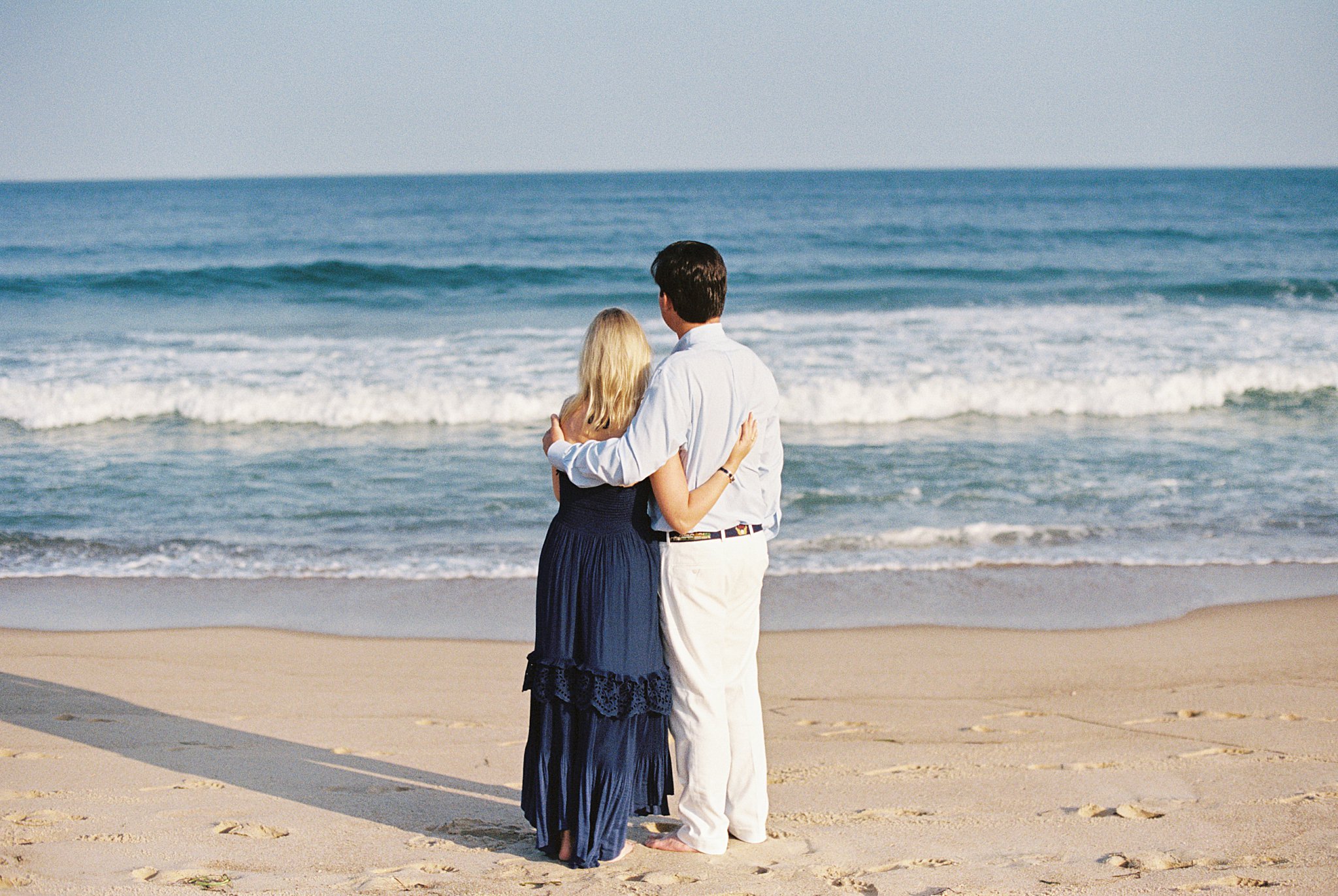 soon to be wed couple looks out to ocean with arms around each other by Lynne Reznick photography 