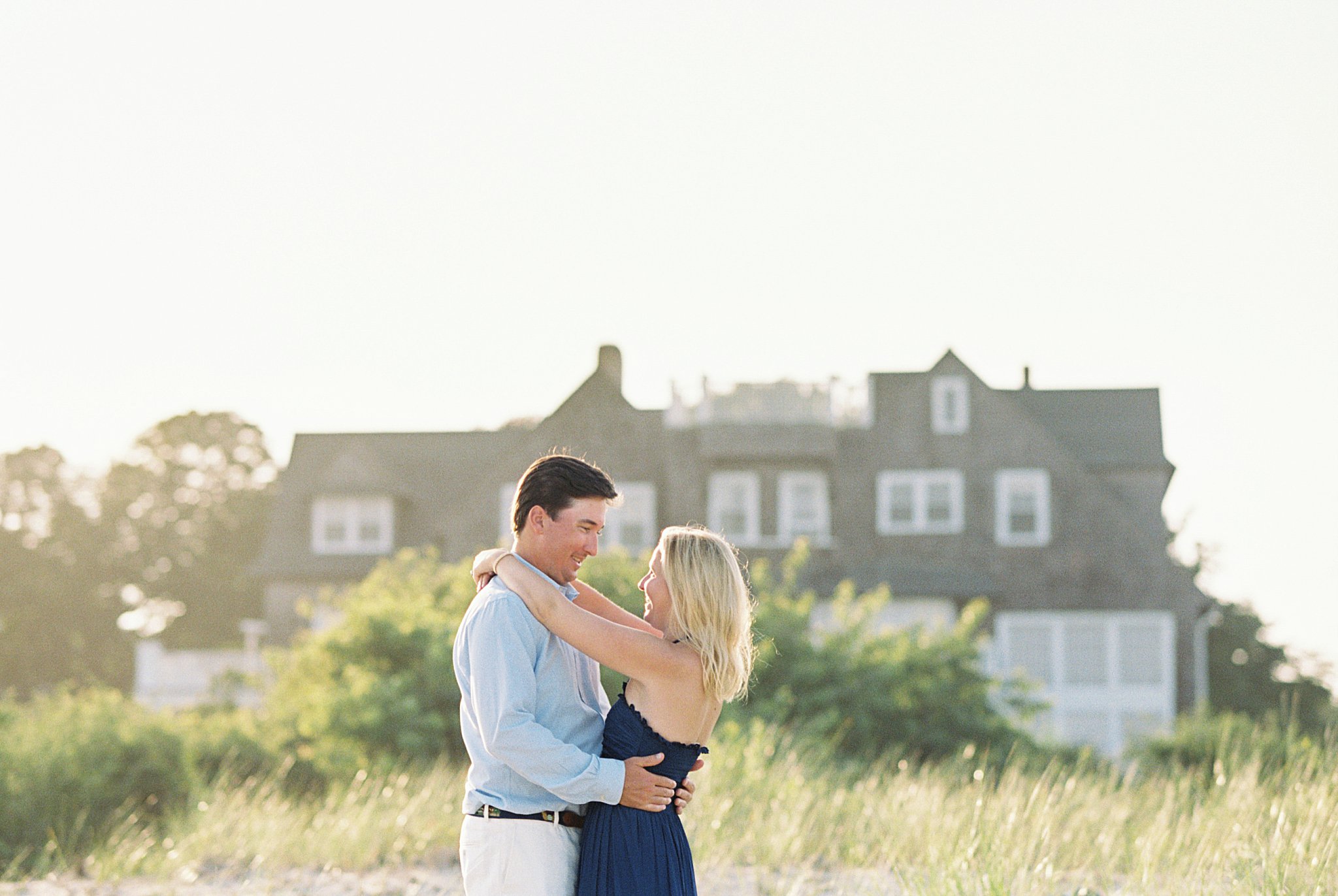 couple wraps arms around each other with house behind them at Watch Hill engagement session