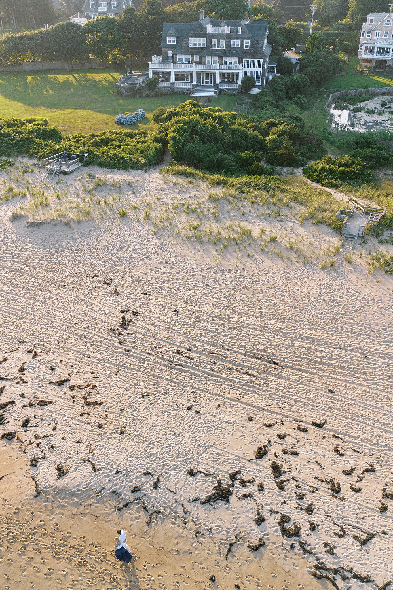 couple walks along beach with family home behind by New England wedding photographer