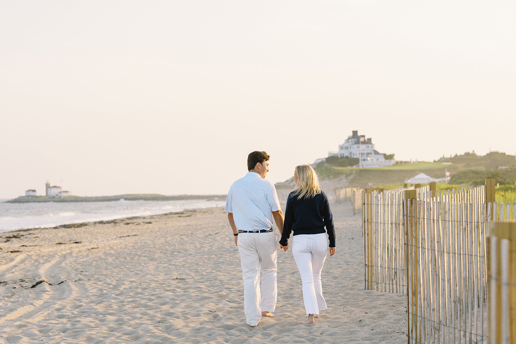 couple walks along sand with lighthouse behind at Watch Hill engagement session