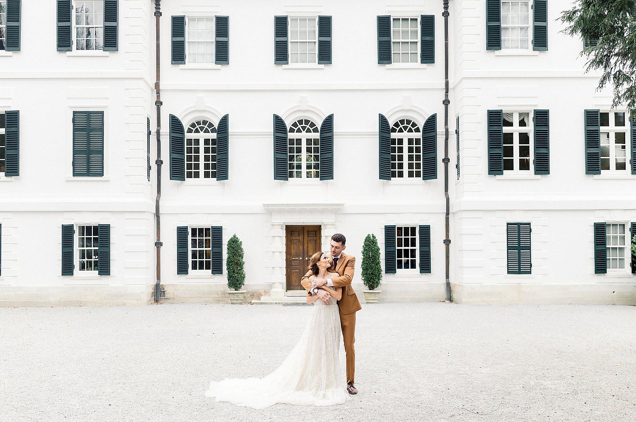 man wraps arms around woman in front of white building with black shutters by New England wedding photographer