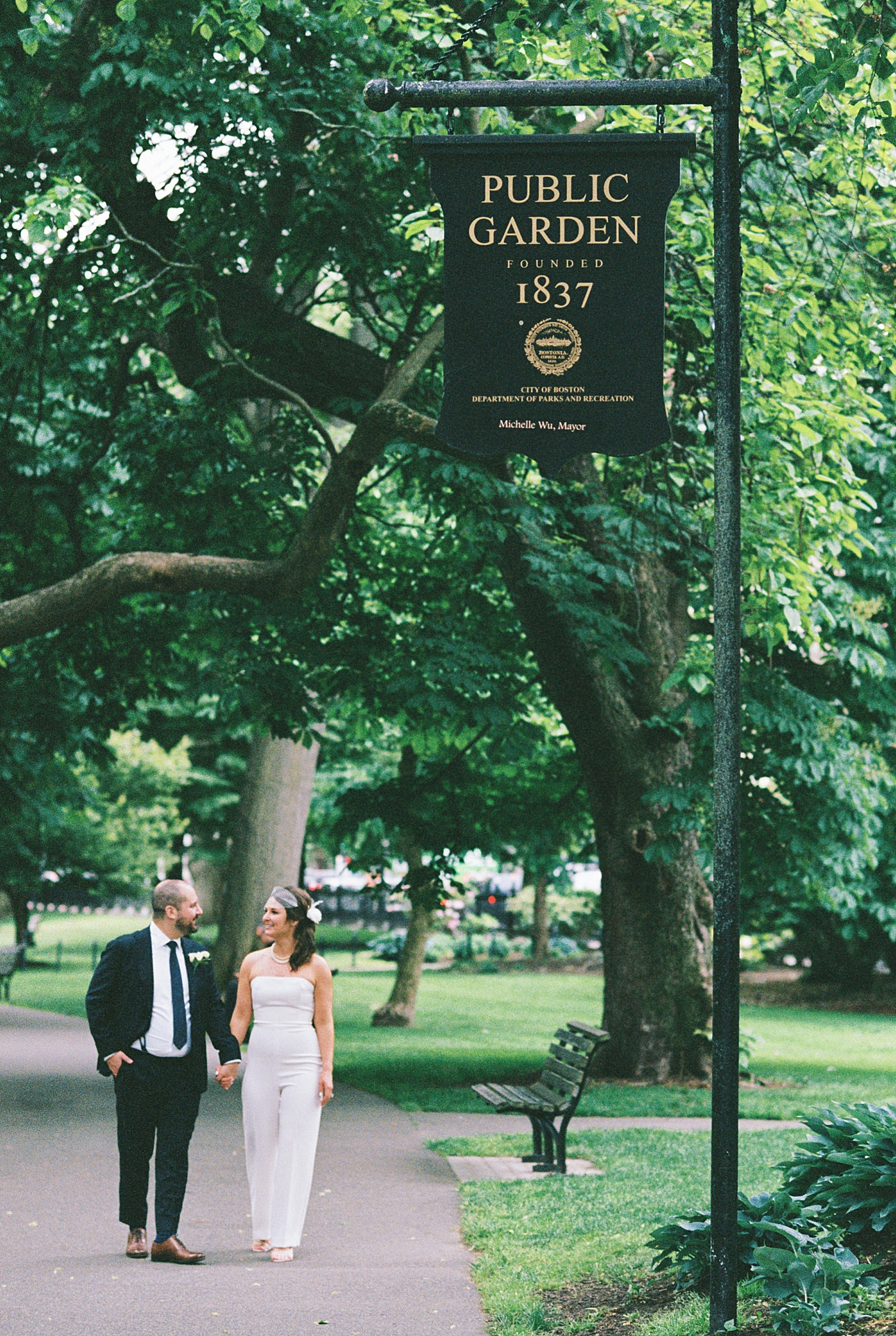 man in suit and woman in white jumper walk under sign for Boston Public Garden