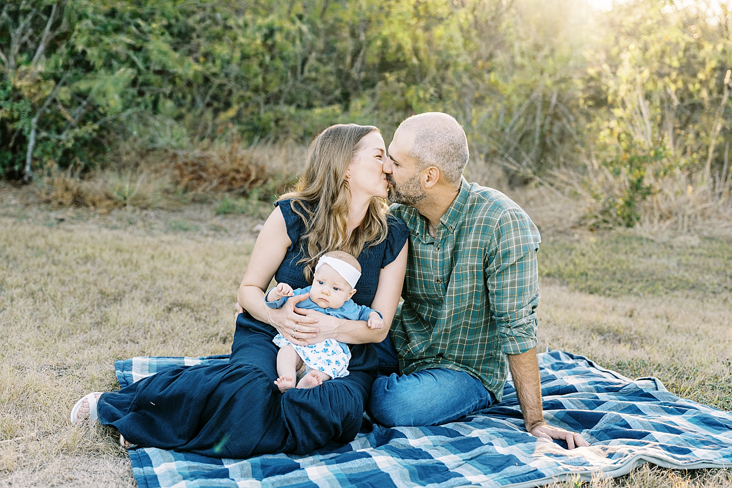 husband and wife share a kiss sitting on blanket holding new daughter by Austin photographer