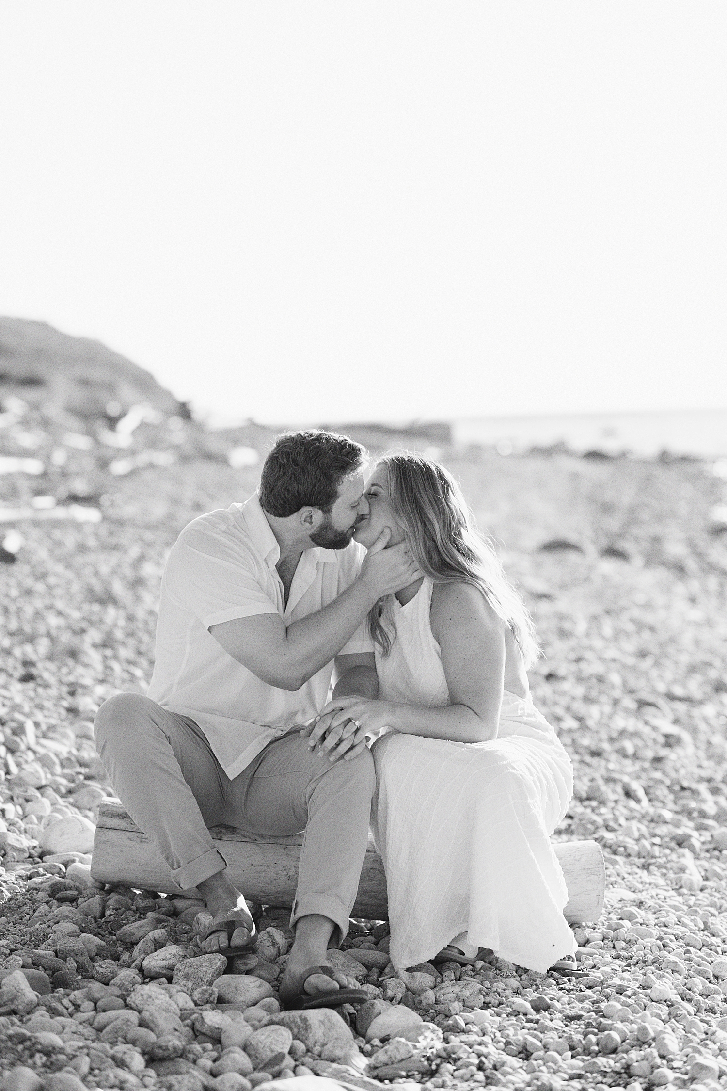 couple sits on driftwood and kisses by the ocean for Montauk engagement session