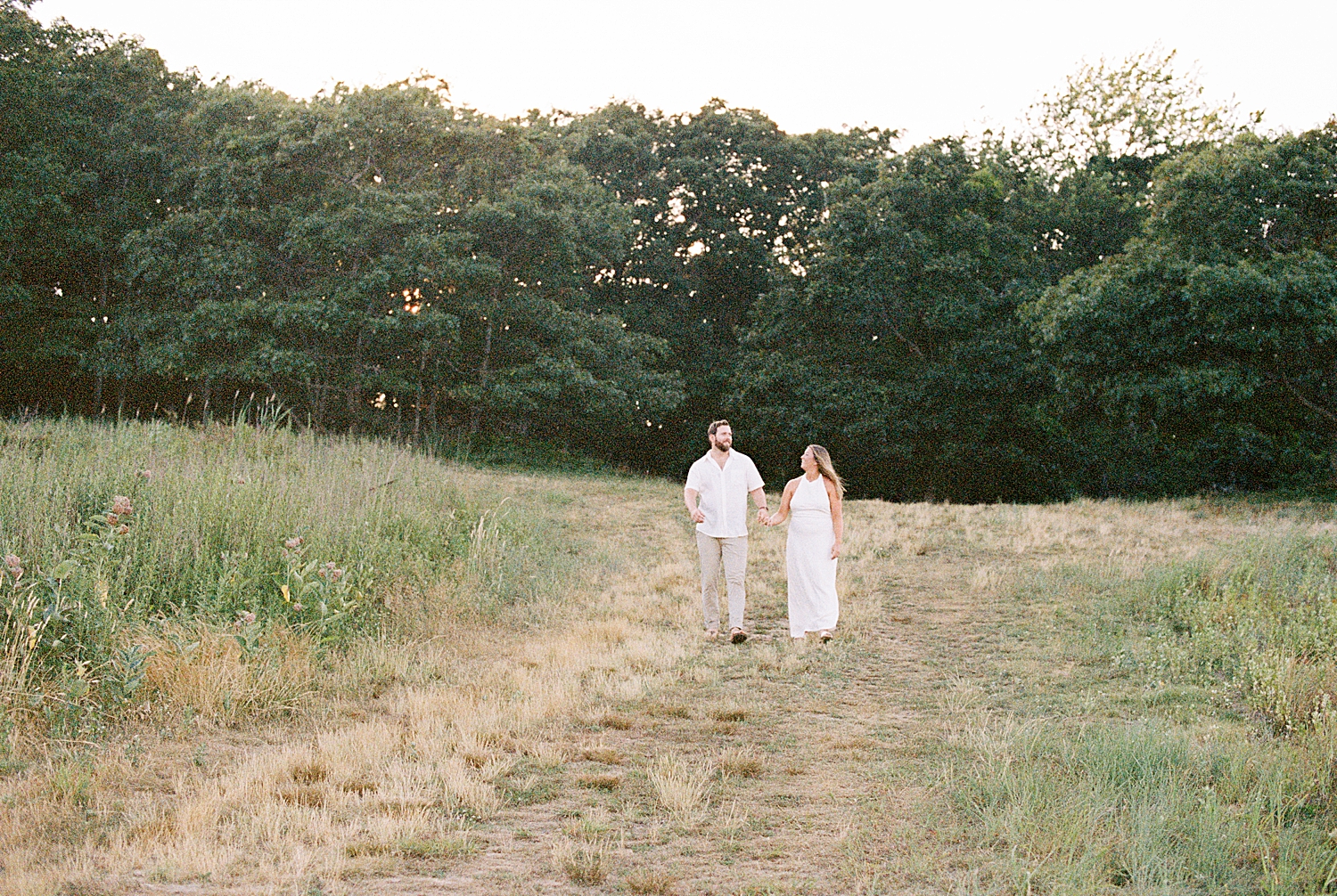 man and woman in white outfits walk through green field for Montauk engagement session