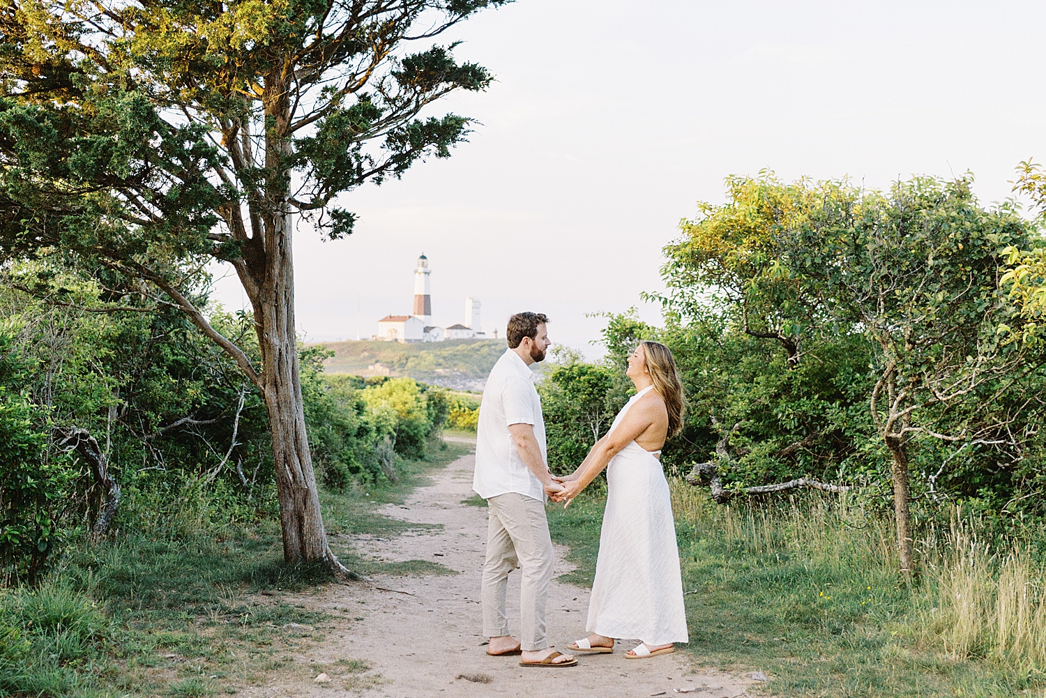 engaged couple hold hands with lighthouse in the distance for Montauk engagement session